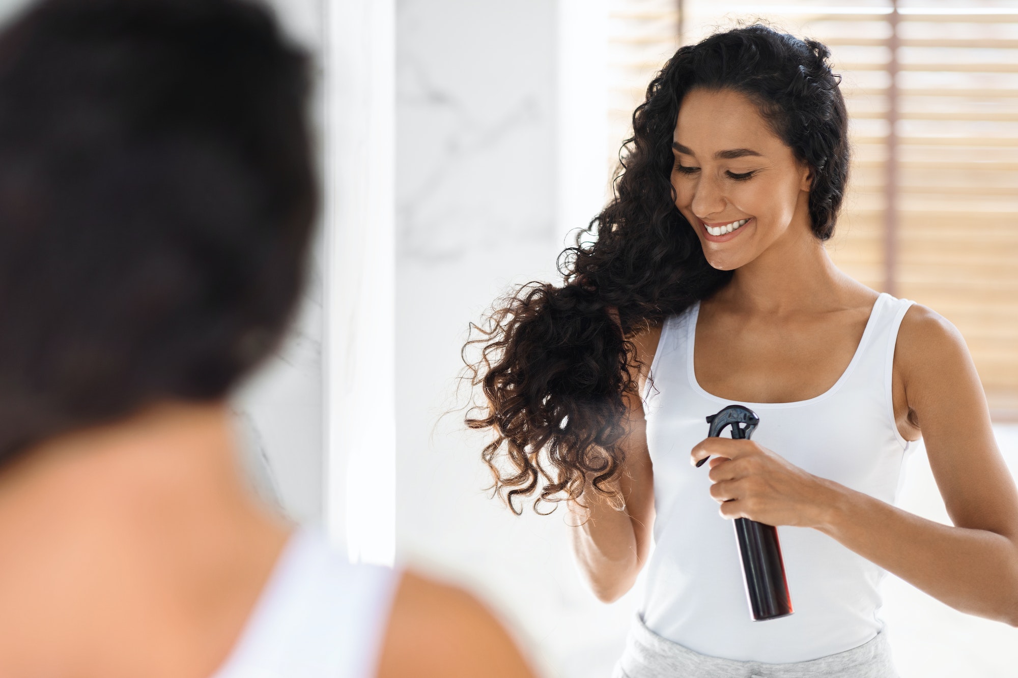 Haircare. Beautiful Young Lady Applying Hair Spray, Standing Near Mirror In Bathroom